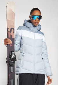 Giacca a vento azzurra chiara con strisce bianche, tasche con zip e cappuccio, abbinata a sci neri e occhiali da sole blu a specchio.