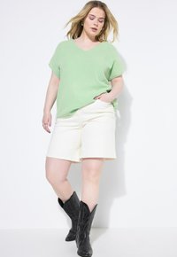 Top de pico texturizado en verde claro combinado con pantalones cortos crema; botas de vaquero negras con detalles acentuados complementan el atuendo.