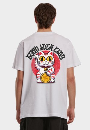 Person iført en hvit t-skjorte med en fargerik maneki-neko-katt og teksten "Good Luck Club" på baksiden mot en ensfarget bakgrunn.