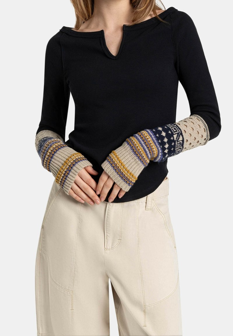 Buste de femme portant un haut noir à manches longues et des chauffe-bras tricotés avec des motifs beige, bleu, jaune et violet, assortis à un pantalon beige clair.