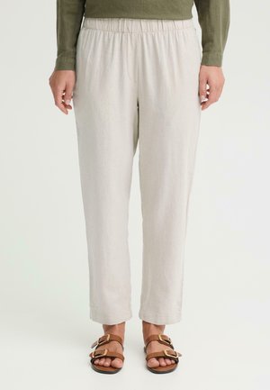 Person iført lyse beige bukser med elastik i taljen og brune sandaler med dobbelte spænder, stående mod en ensfarvet lys baggrund.