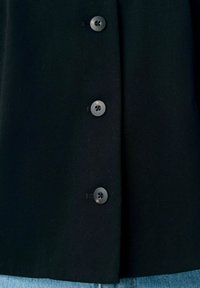 Camicia o giacca nera con tre bottoni scuri visibili sopra jeans in denim azzurro chiaro.