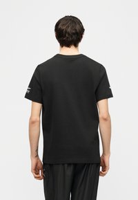 Άτομο με κοντά καστανά μαλλιά στέκεται με την πλάτη προς το μέρος μας, φοράει απλό μαύρο κοντομάνικο μπλουζάκι και μαύρο παντελόνι, μπροστά σε φωτεινό φόντο.