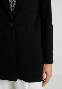 Černý strukturovaný blazer s jedním knoflíkem, dvěma předními kapsami a vypasovaným střihem, zobrazený částečně s bílým oděvem pod ním.