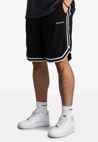 Shorts noirs avec des rayures blanches sur les côtés et le logo "PEGADOR". Fabriqués en tissu doux, associés à des baskets blanches et des chaussettes rayées.