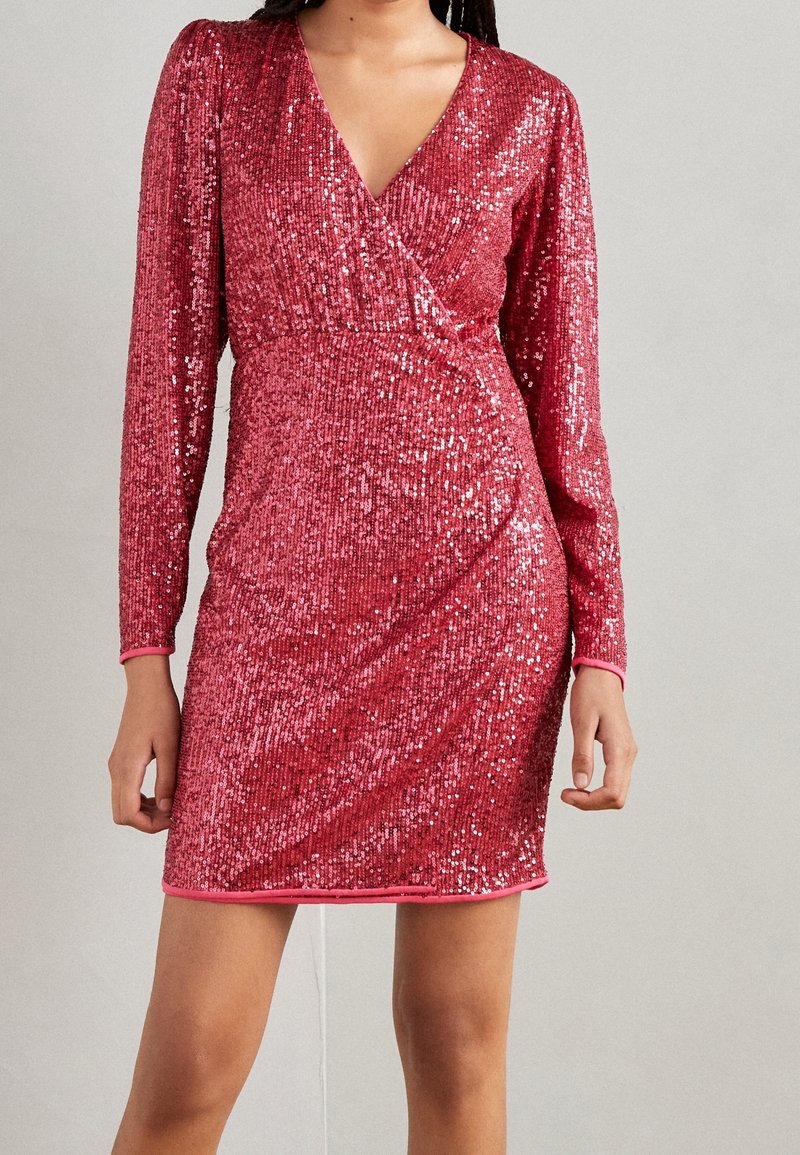 Kvinna klädd i en långärmad, rosa paljettminiklänning med V-ringning och omslagstyp framtill mot en enkel bakgrund.