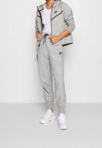 Ljust grå zip-up hoodie med svarta detaljer, kombinerad med matchande grå joggers med dragsko i midjan och sidofickor, bärs med vita sneakers.