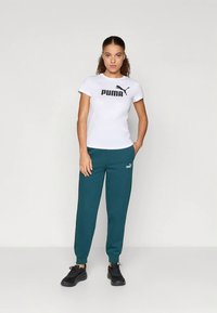 Γυναίκα που στέκεται, φορώντας λευκό t-shirt Puma, τιρκουάζ φόρμες Puma και μαύρα αθλητικά παπούτσια, σε απλή φωτεινή φόντο.