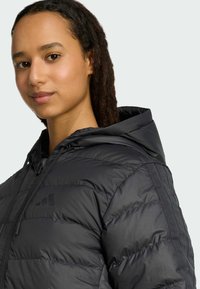 Crna pufasta jakna s kapuljačom, koja se ističe prošivanim dizajnom i teksturiranim panelima. Uključuje mali, crni logo na prsima i vezicu oko vrata.
