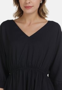 Schwarzes Kleid mit V-Ausschnitt, gerafftem Bund und langen Ärmeln. Glattes Material mit leichtem Schimmer, mit elastischen Akzenten an den Ärmeln.