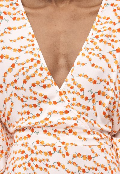 Κοντινό πλάνο ενός φόρεματος τύπου wrap με βαθύ V-λαιμόκοψη, με λευκό φόντο και πορτοκαλί λουλουδένιο σχέδιο.