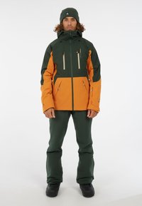 Wasserdichte Jacke in Dunkelgrün und Orange, mit Reißverschluss, Seitentaschen und hohem Kragen. Kombiniert mit passenden grünen Hosen und schwarzen Stiefeln.