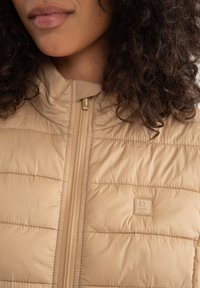 Béžový nadýchaný vest s vysokým límcem, prošívanou texturou a zlatým zipem. Na levé straně se nachází značková nášivka. Měkký, lehký materiál.