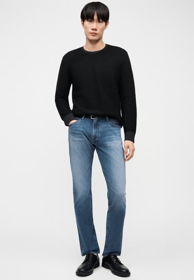 Νέος άνδρας που φοράει μαύρο υφασμάτινο πουλόβερ, μπλε τζιν με ζώνη και μαύρα δερμάτινα παπούτσια, στέκεται μπροστά σε απλό λευκό φόντο.