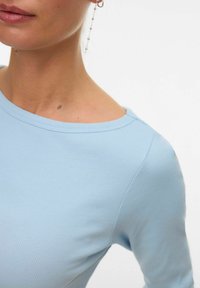 Close-up van een persoon die een lichtblauw geribd shirt draagt en een lange zilveren oorbel met kleine kralen.