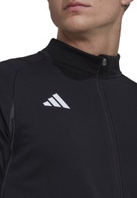 Schwarze Sportjacke mit hohem Kragen, Reißverschluss und weißem Logo auf der Brust. Sie weist eine glatte Textur und eine taillierte Passform auf.