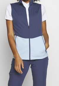 Navy and light blue sleeveless vest with a high collar, zippered front, and hand pockets; fabric appears smooth and lightweight.