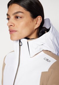 Weiße und beige isolierte Jacke mit hohem Kragen, mit einem Reißverschluss vorne und CMP-Logo auf der Schulter. Weicher, strukturiertes Gewebe.
