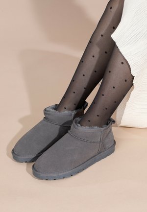 Bottines en suède gris avec une doublure en fourrure douce et des accents cousus, sur un fond beige, portées avec des collants noirs à pois.
