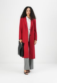 Long manteau rouge avec col cranté, deux boutons et poches latérales. Associé à une chemise légère, un pantalon gris et un sac à main noir.