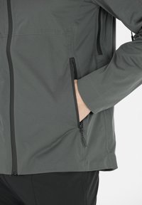 Grijze zip-up jas met een gladde textuur, voorzien van zijzakken met zwarte ritsen en een getailleerd ontwerp. Geen zichtbare logo's of patronen.
