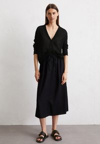 Schwarze Strickjacke mit V-Ausschnitt und Knöpfen, kombiniert mit einem marineblauen Midirock mit Kordelzugbund. Getragen mit schwarzen flachen Sandalen.