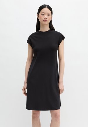 Młoda kobieta z długimi czarnymi włosami, ubrana w czarną, bezrękawnikową sukienkę do kolan, stoi na jednolitym białym tle.