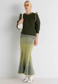 Oliwkowy sweter dzianinowy z falbaniastymi akcentami, połączony z żółtą spódnicą maxi w ombre, z pionowymi pliskami i ząbkowanym hemem, noszoną z białymi butami.