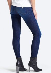 Vaqueros skinny de mezclilla oscura con una textura suave, que presentan dos bolsillos traseros y un detalle del logo triangular cerca de la cintura. Llevados con botas de tacón negras.
