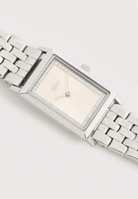 Montre-bracelet rectangulaire argentée avec un cadran rose pâle bordé de cristaux et un logo crocodile argenté à 12 heures.