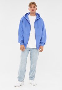 Felpa con zip di colore azzurro chiaro, con cappuccio regolabile e due tasche anteriori, indossata sopra una maglietta bianca, abbinata a jeans in denim chiaro e sneakers bianche.