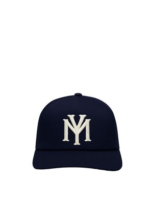 Jeune homme aux yeux clairs portant une casquette et une chemise bleu marine, toutes deux avec des logos blancs brodés, regardant légèrement sur le côté.
