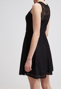 Schwarzes Spitzekleid mit tailliertem Oberteil und ausgestelltem Rock. Verfügt über eine floral gemusterte Textur und ärmelloses Design. Der Rücken zeigt Spitzendetails.