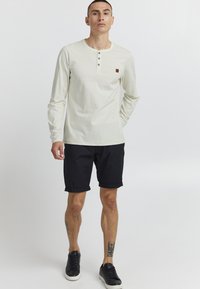 Camisa de manga larga color crema con tapeta de botones, combinada con pantalones cortos negros enrollados y zapatillas negras. Presenta un pequeño parche de logo marrón.