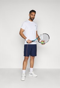 T-shirt bianco a maniche corte con logo verde, pantaloni corti blu navy con piping arancione e sneaker bianche. Tiene in mano una racchetta da tennis e una pallina.