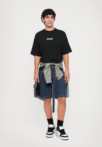 T-shirt nera con logo "BARROW", gonna di jeans blu legata con una camicia a quadri, sneaker nere e una borsa nera piccola.