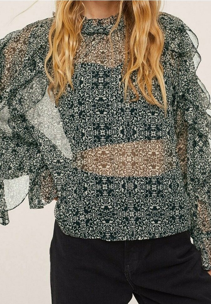 Femme portant un chemisier transparent à fleurs noir et blanc avec de longues manches à volants et un pantalon noir.