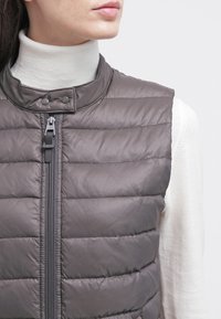 Gilet grigio imbottito con colletto alto, zip frontale completa e bottoni a pressione, indossato sopra un dolcevita crema. Texture liscia e leggera.