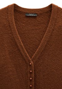 Brązowy, dziany kardigan z dekoltem w kształcie litery V. Posiada pięć brązowych guzików i miękką, teksturowaną tkaninę. Metka widoczna przy dekolcie.