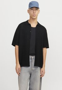 Cardigan corto a maniche corte in maglia nera con un motivo testurizzato e chiusura a bottoni, indossato sopra un top a costine, abbinato a jeans in denim chiaro e un cappellino blu.