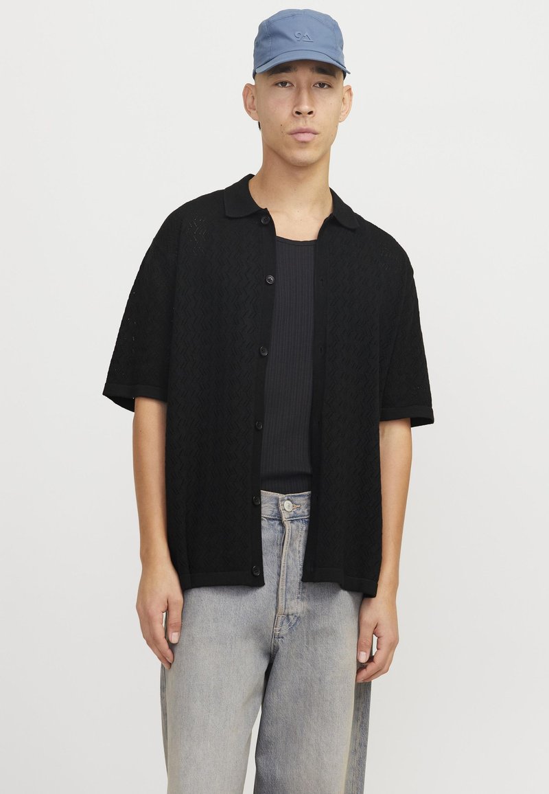 Cardigan corto a maniche corte in maglia nera con un motivo testurizzato e chiusura a bottoni, indossato sopra un top a costine, abbinato a jeans in denim chiaro e un cappellino blu.