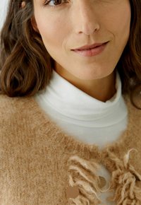 Beige fuzzy cardigan met een zachte textuur en subtiele franjes. Witte coltrui eronder, met een gesloten halslijn.