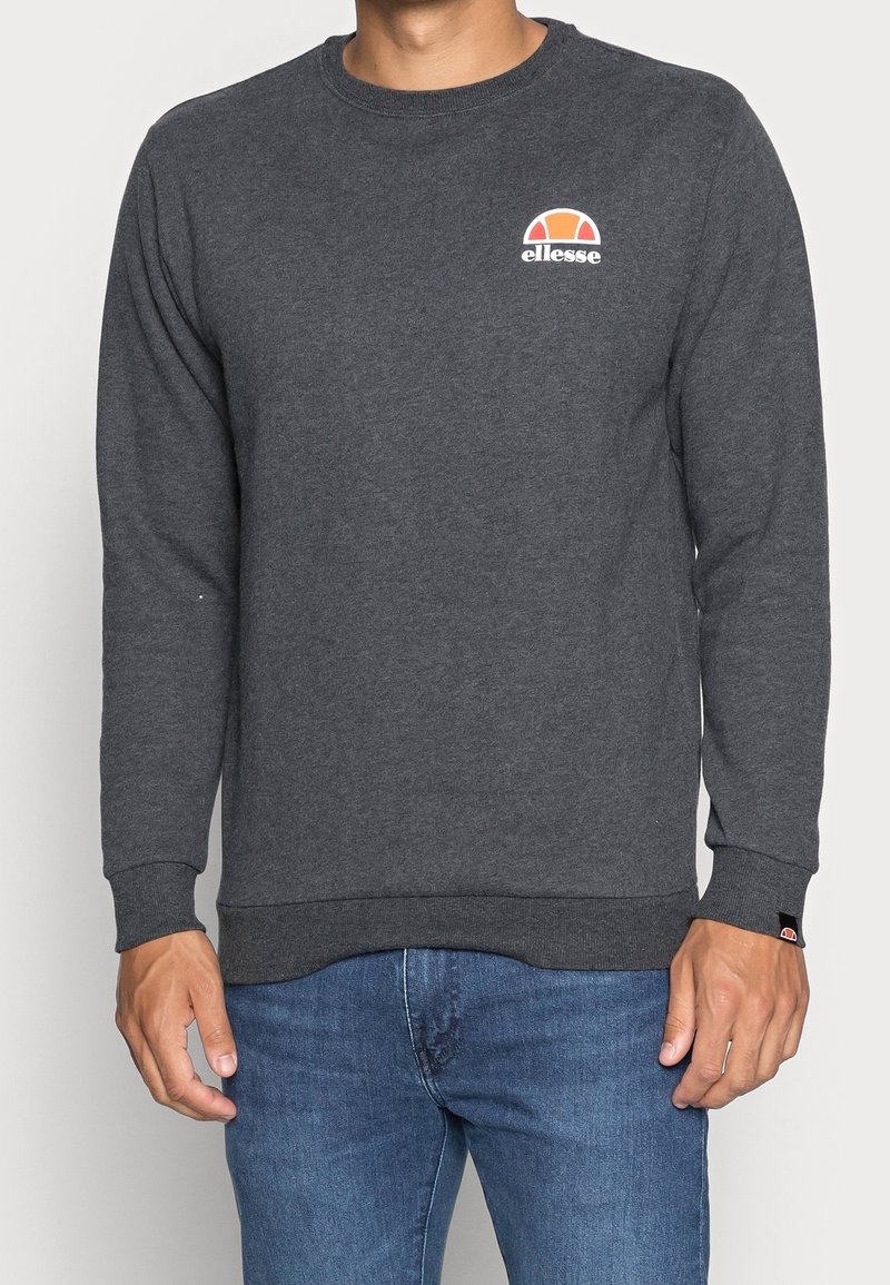 Homme portant un sweat-shirt gris foncé à col rond avec un petit logo ellesse orange et blanc sur la poitrine et un jean bleu.