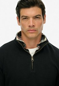 Czarny sweter z dzianiny z zamkiem przy kołnierzu. Wewnętrzna część kołnierza ma wzór w kratę w odcieniach czerwieni i zieleni. Detale obejmują małe logo na piersi.