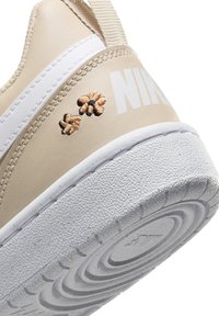 Bežna športna obutev z teksturirano površino, belimi poudarki in odtisnjenim logotipom. Dekorativna vezenina rjavih cvetov blizu pete. Gumijast podplat.