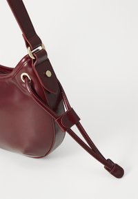 Red leather shoulder bag with a curved shape, gold hardware accents, and a sleek adjustable strap. Smooth texture and minimalist design.