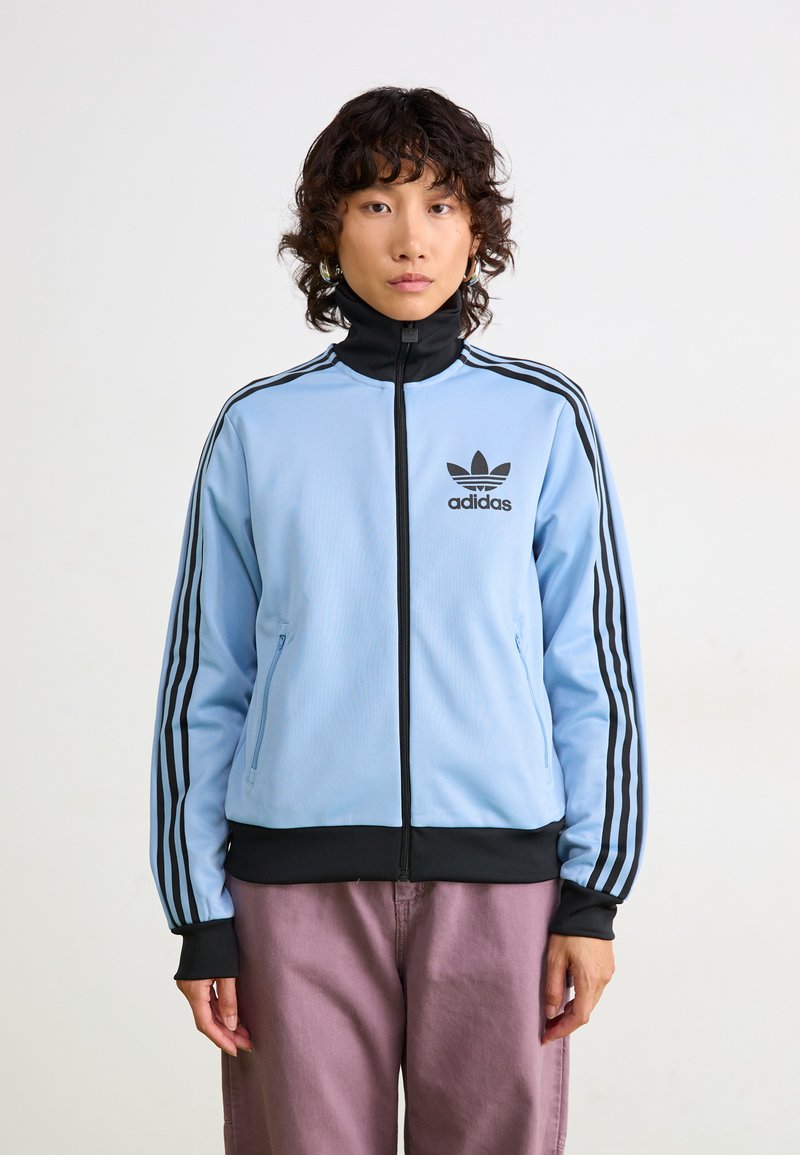 Asmuo, vilkintis šviesiai mėlyną „Adidas“ treniruočių švarką su juodomis juostelėmis ir logotipu, derinantis su mauve spalvos kelnėmis, stovintis prieš paprastą foną.