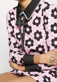 Rosa klänning med svart hexagonmönster, svart krage med pärldetaljering, knytband vid halsen, långa ärmar med svarta manschetter.