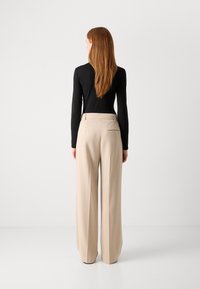 Bež hlače širokog kroja s glatkom teksturom, plisiranim detaljima i jednim stražnjim džepom. Crni uski top s dugim rukavima vidljiv sa stražnje strane.