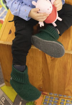 Green felt slippers with ribbed cuffs, gray soles, worn by a seated child holding a pink stuffed toy, on a wooden surface.